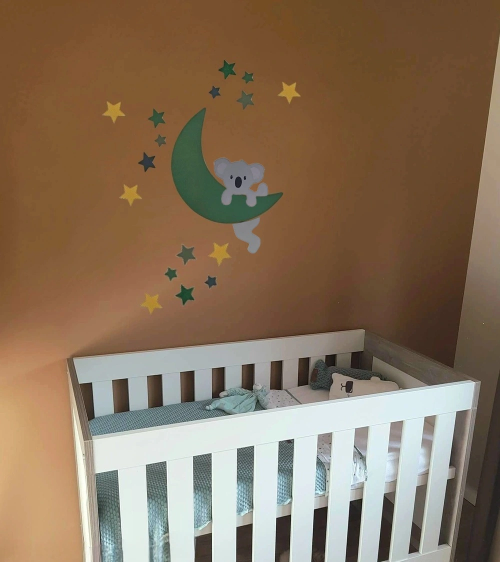 Behangsticker met een hangend koalabeertje aan een maan met sterren in donkergroen en oker op een bruine wand in de kinderkamer.